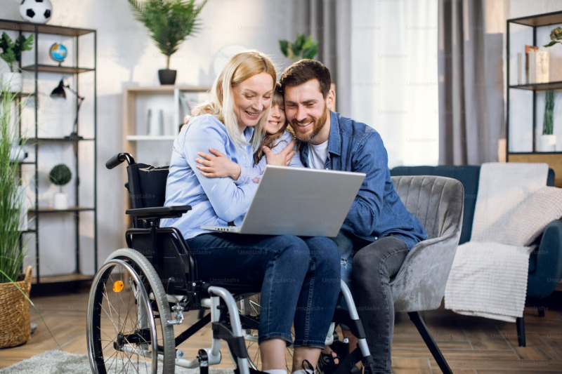 Wife in a wheelchair hugging husband and child. Wife has laptop on knees.